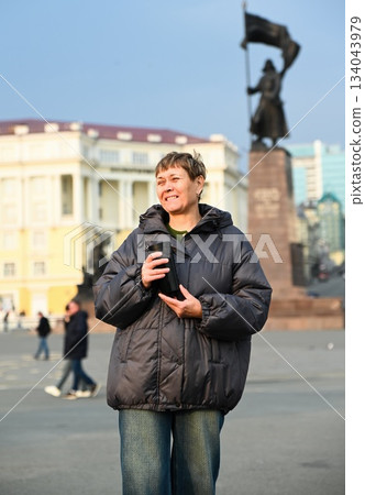 Russian woman 60 years old stands on city square in Vladivostok holding a thermal mug of coffee enjoying calm moment and morning mindfulness and peace Russian woman 60 years old stands on city square in Vladivostok holding a thermal mug of coffee enjoying calm moment and morning mindfulness and peace 134043979