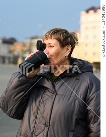 Russian woman 60 years old drinks coffee from a thermal mug outdoors in Vladivostok enjoying warm sunlight calm morning mood and simple mindful moment 134043980