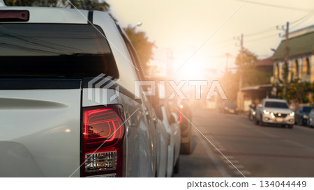 Side of pickup car parking beside road. Dim light of a warm evening. Other car driving on clear road. City and buildings along the road. Under light and evening sky. 134044449