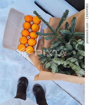 Winter overhead view of snow boots next to a wrapped fir branch and carton of fresh oranges on snowy ground. Winter overhead view of snow boots next to a wrapped fir branch and carton of fresh oranges on snowy ground. 134044963
