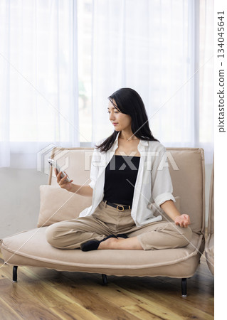 A woman meditating on a sofa while looking at her smartphone A woman meditating on a sofa while looking at her smartphone 134045641