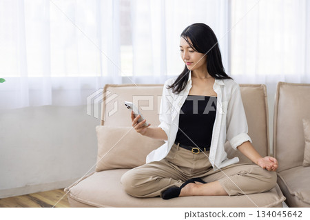 A woman meditating on a sofa while looking at her smartphone A woman meditating on a sofa while looking at her smartphone 134045642