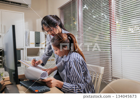 Young business women team work online, home office people collaborate learn, reviewing documents near computer desk, standing colleague writing on clipboard, seated woman holds large paper roll. Young business women team work online, home office people collaborate learn, reviewing documents near computer desk, standing colleague writing on clipboard, seated woman holds large paper roll. 134045673