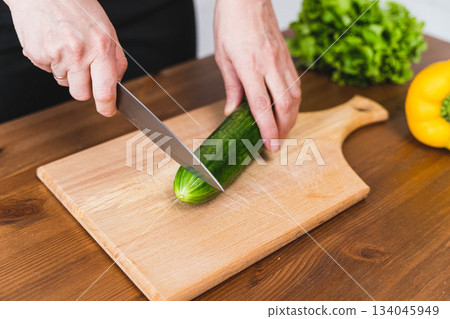 Person cuts cucumber on wooden board in kitchen during meal preparation Person cuts cucumber on wooden board in kitchen during meal preparation 134045949