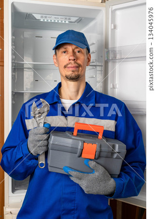 Repair technician holds toolbox and wrench in front of empty refrigerator during appliance service Repair technician holds toolbox and wrench in front of empty refrigerator during appliance service 134045976