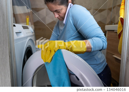 Woman cleaning washing machine while talking on phone in laundry room Woman cleaning washing machine while talking on phone in laundry room 134046016