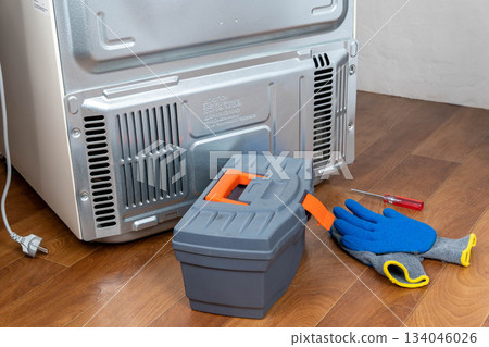 Repair tools and gloves are near the back of a refrigerator on a wooden floor Repair tools and gloves are near the back of a refrigerator on a wooden floor 134046026