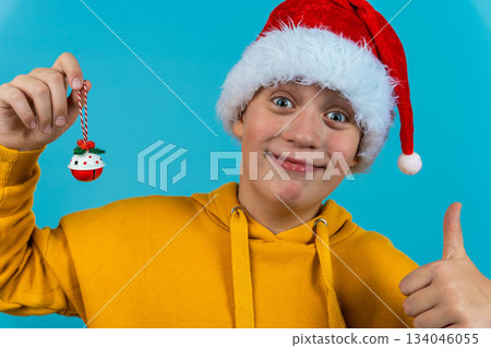 Boy in a yellow hoodie shows a Christmas ornament while wearing a Santa hat Boy in a yellow hoodie shows a Christmas ornament while wearing a Santa hat 134046055