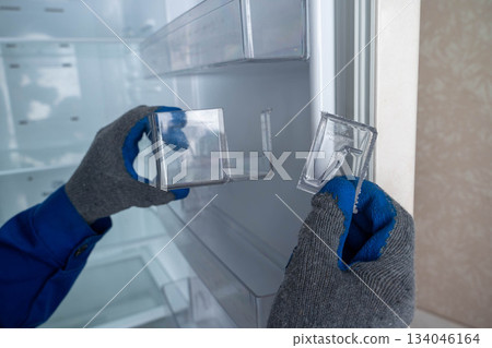 The hands of a home appliance maintenance technician inspects a broken plastic shelf from the door of a home refrigerator 134046164