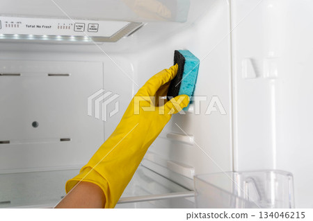 Cleaning a refrigerator with a sponge while wearing yellow gloves in a kitchen Cleaning a refrigerator with a sponge while wearing yellow gloves in a kitchen 134046215