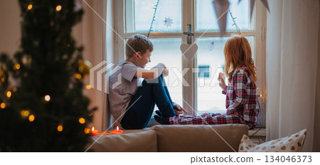 Small girl and boy sitting on window sill in pajamas. Christmas time. Small girl and boy sitting on window sill in pajamas. Christmas time. 134046373