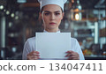 Caucasian female chef holding up a white piece of paper with a serious face 134047411