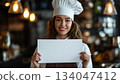 A foreign female chef smiling and holding up a white piece of paper 134047412