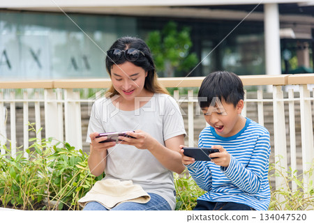 Mother, child, summer city scene, family woman and son enjoy playing mobile games outdoors, parent and child connecting with technology on urban street, people having fun. 134047520