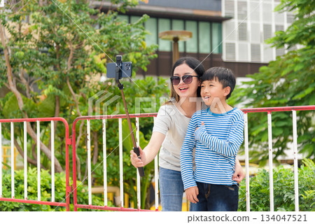 Mother, child, parent, son, happy family people, woman enjoys summer city street outdoor activity, capturing cheerful moment with selfie stick, takes mobile phone picture, urban street setting. Mother, child, parent, son, happy family people, woman enjoys summer city street outdoor activity, capturing cheerful moment with selfie stick, takes mobile phone picture, urban street setting. 134047521