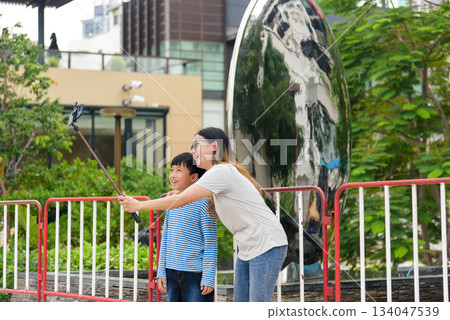 Mother, son, child, family, woman, parent, people find summer joy on city street, taking cheerful selfie outdoors, urban background shows vibrant foliage, reflective buildings. 134047539