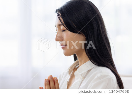 A woman meditating by the window with her hands together and her eyes closed A woman meditating by the window with her hands together and her eyes closed 134047698