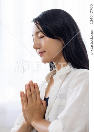 A woman meditating by the window with her hands together and her eyes closed A woman meditating by the window with her hands together and her eyes closed 134047700