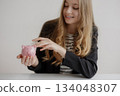 Young woman smiling while holding a pink piggy bank in a modern indoor setting during daylight 134048307