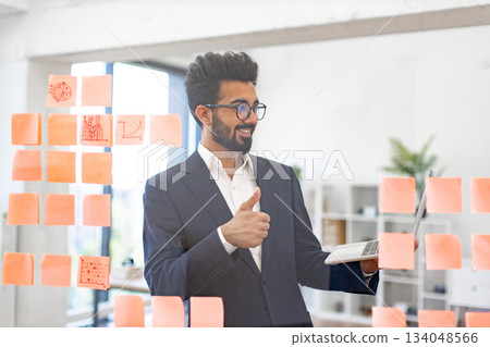 An Indian businessman smiles while holding a laptop and giving a thumbs up, showing approval An Indian businessman smiles while holding a laptop and giving a thumbs up, showing approval 134048566