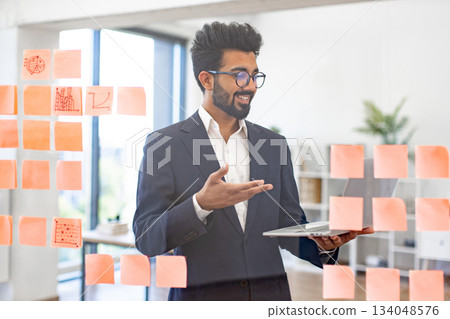 An Indian businessman smiles while presenting his ideas on sticky notes with a laptop in hand 134048576