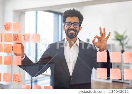 An Indian businessman smiles while making an okay gesture, working on a project with sticky notes An Indian businessman smiles while making an okay gesture, working on a project with sticky notes 134048611