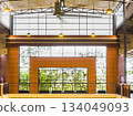 Industrial interior with exposed steel trusses, brick wall detailing, and large grid windows framing lush greenery. Clean lines, warm lighting, and a balanced architectural composition. 134049093