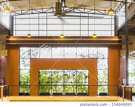 Industrial interior with exposed steel trusses, brick wall detailing, and large grid windows framing lush greenery. Clean lines, warm lighting, and a balanced architectural composition. 134049093