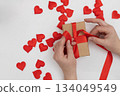 Person Tying a Red Ribbon Around a Gift Box Surrounded by Red Hearts for Valentine's Day 134049549