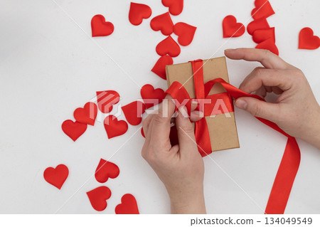 Person Tying a Red Ribbon Around a Gift Box Surrounded by Red Hearts for Valentine's Day Person Tying a Red Ribbon Around a Gift Box Surrounded by Red Hearts for Valentine's Day 134049549