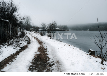 Lonely Dirt Road Curving Along a Snowy Riverbank in Winter Fog 134050402
