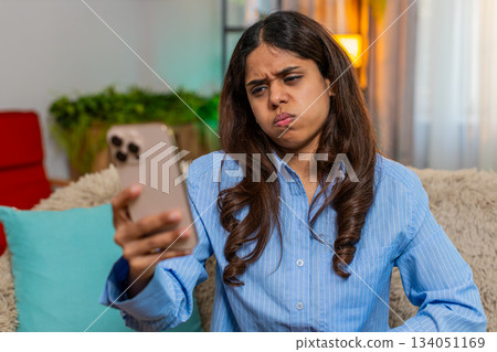 Indian woman looking down with sad sigh while reading bad news on smartphone on home sofa indoors 134051169