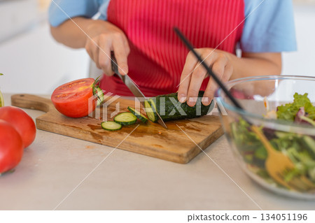 Young woman in kitchen wearing apron slices cucumber on board with tomatoes makes fresh salad bowl 134051196