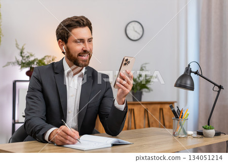 Middle-aged businessman on smartphone video call conversation wears earphones writes notes smiling Middle-aged businessman on smartphone video call conversation wears earphones writes notes smiling 134051214