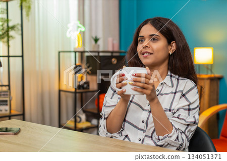 Indian woman enjoying hot coffee eyes closed holding cup savoring aroma relaxing peacefully indoors Indian woman enjoying hot coffee eyes closed holding cup savoring aroma relaxing peacefully indoors 134051371