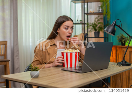 Young woman watching dramatic movie on laptop eating popcorn nervously reacting to intense scene Young woman watching dramatic movie on laptop eating popcorn nervously reacting to intense scene 134051439