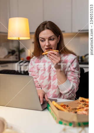 Adult woman works on laptop eats pizza at night enjoys hot slice smiles cozy mood happy productivity Adult woman works on laptop eats pizza at night enjoys hot slice smiles cozy mood happy productivity 134051553