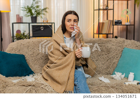 Young woman wrapped in plaid blanket sitting on sofa holding hot tea feeling sick with cold symptoms Young woman wrapped in plaid blanket sitting on sofa holding hot tea feeling sick with cold symptoms 134051671