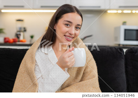 Young woman drinks tea wrapped in warm blanket relax calm cozy rest soft smile gentle mood slow pace 134051946