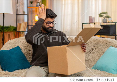 Indian man on sofa opens box and shakes head after discovering wrong item in online delivery order 134051951