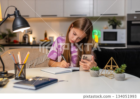 Teenager girl online math lesson on smartphone serious focus writes notes in notebook for schoolwork 134051993