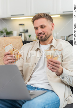 Young man counts cash euros compares with laptop earnings sheet fills sums smiles proud at goal met Young man counts cash euros compares with laptop earnings sheet fills sums smiles proud at goal met 134052003