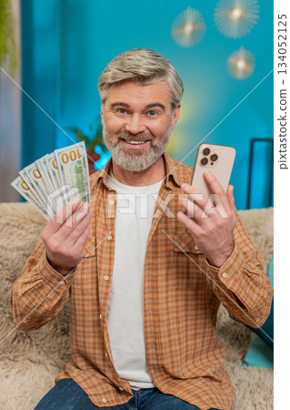 Mature man with smartphone and money, making financial transfer via mobile app, smiling at home Mature man with smartphone and money, making financial transfer via mobile app, smiling at home 134052125