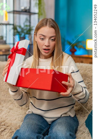 Caucasian woman opening birthday surprise gift on sofa smiling excited indoors happy joyful reaction 134052249