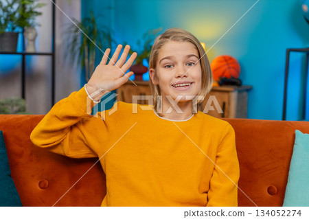 Teenager boy waving with smile greeting happily sitting on sofa, looking straight at camera at home 134052274