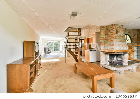 A stylish and cozy modern living room featuring a staircase, wooden furniture, a fireplace, and large windows for natural light. 134052637