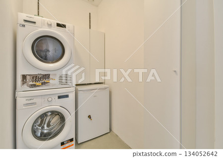 A contemporary laundry room featuring stacked washing machine and dryer, along with a compact refrigerator. The simplicity and cleanliness of the space highlight modern design. A contemporary laundry room featuring stacked washing machine and dryer, along with a compact refrigerator. The simplicity and cleanliness of the space highlight modern design. 134052642