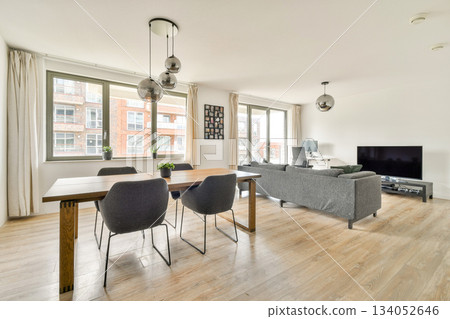 A stylish and contemporary living room featuring a wooden dining table, comfortable seating, and large windows allowing natural light into the space. 134052646