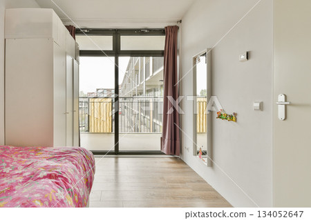 A stylish modern bedroom featuring a pink floral bedspread, large windows, and a sliding door that opens to a balcony overlooking city architecture. 134052647
