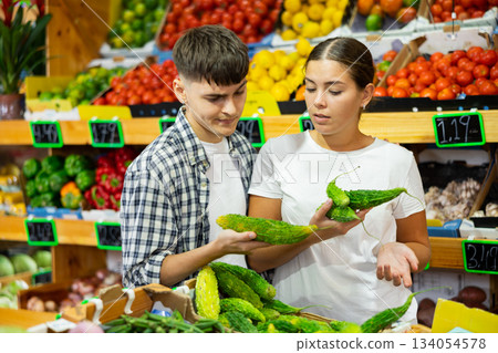 Positive couple standing together in fruit and vegetable section of supermarket and choosing indian cucumber momordica Positive couple standing together in fruit and vegetable section of supermarket and choosing indian cucumber momordica 134054578